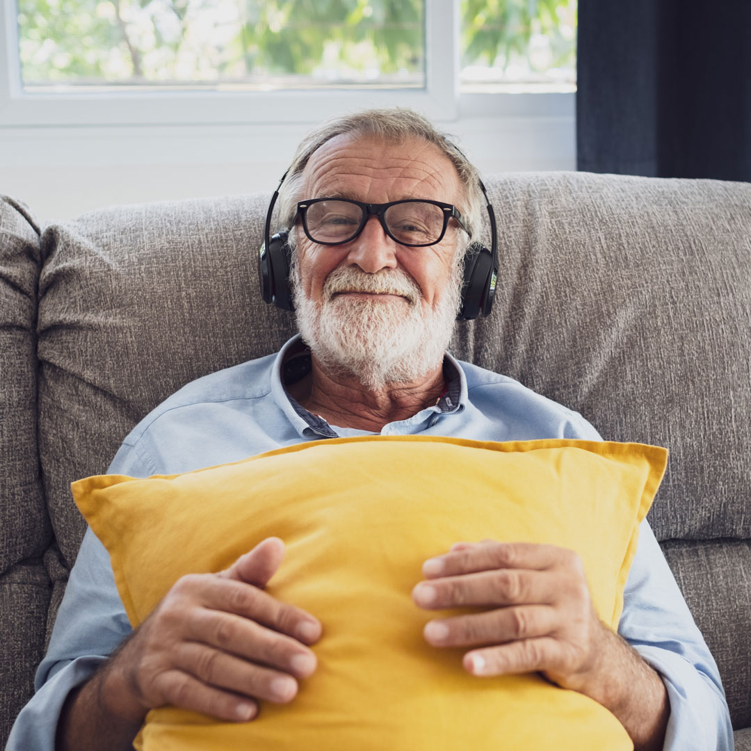 Ein älterer Herr mit weiß-grauem Haar und Bart sitzt auf einem Sofa, trägt Kopfhörer und lächelt mit einem gelben Kissen in den Händen in die Kamera.