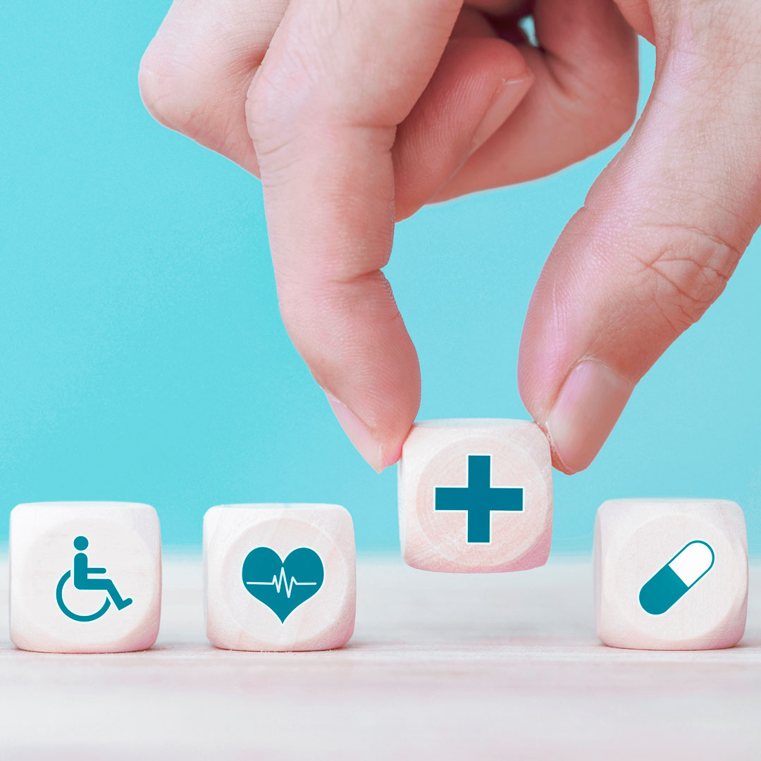 Vier Würfel zeigen verschiedene Symbole aus dem medizinischen Bereich. Eine Hand hebt den Würfel mit einem Ambulanzkreuz an.